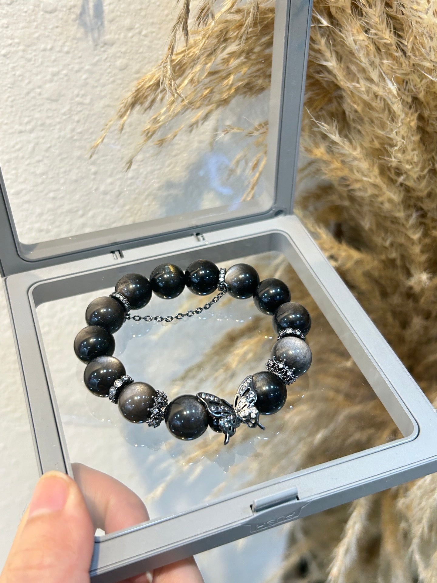 Silver obsidian bracelet with black butterfly charm, photographed in a clear display box against a natural pampas background – for protection, vitality, and style.