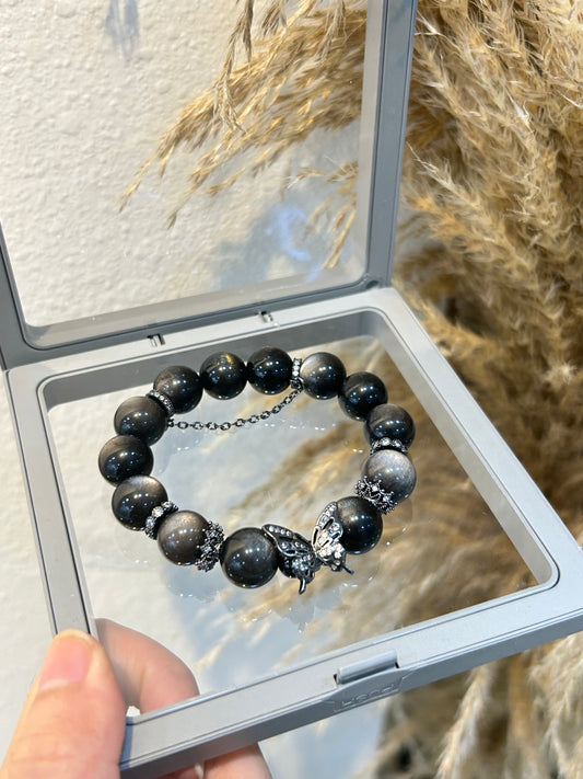 Silver obsidian bracelet with black butterfly charm, photographed in a clear display box against a natural pampas background – for protection, vitality, and style.