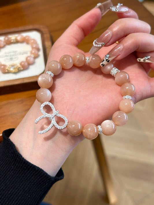 Hand holding peach moonstone bracelet with rhinestone bow charm – symbolizing fortune, elegance, and emotional renewal.