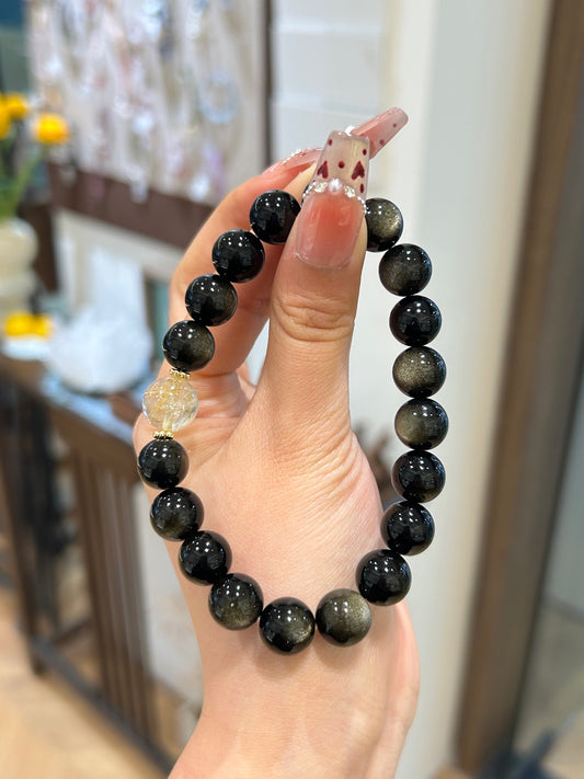 Hand holding gold sheen obsidian bracelet with single golden rutilated quartz bead – symbolizing protection, clarity, and focused wealth energy.